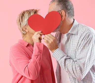 Visuel d'un couple se faisant un bisous caché derrière un cœur rouge en carton
