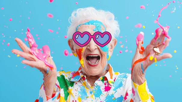 Personne âgé souriante avec des lunettes un cœur dessiné sur le visage et levant des mains pleines de peinture