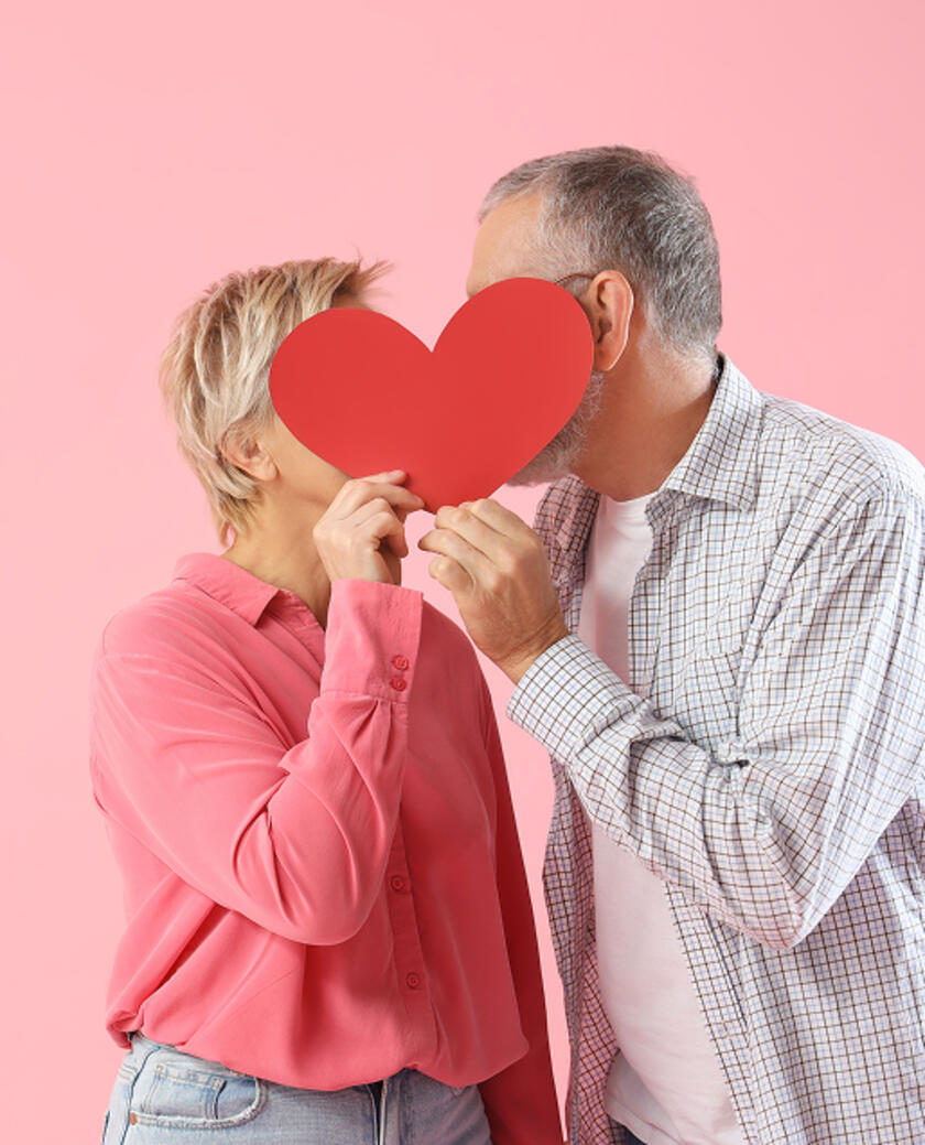 Visuel d'un couple se faisant un bisous caché derrière un cœur rouge en carton